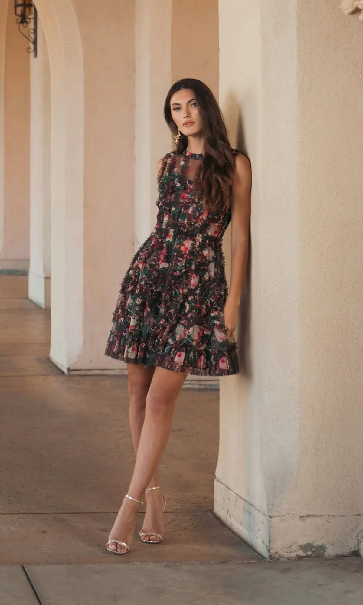 A woman in a High-Neck Print Wedding-Guest Dress, Mac Duggal 8307, with floral pattern and heels stands by a light-colored wall with arches in the background.